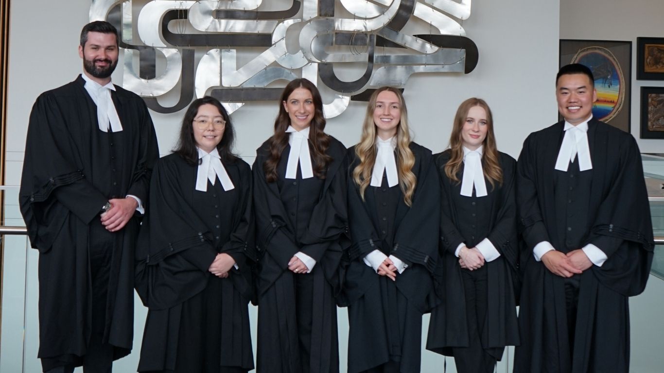 Six newly called TDS law associates, smiling and wearing traditional black legal robes and white tabs, standing in a row for a group photo in front of a silver wall sculpture.